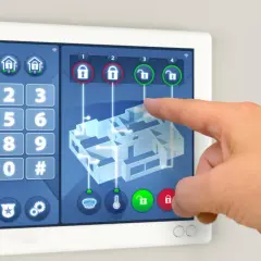 Palec dotyka ekranu panelu alarmowego, pokazując schemat domu. Czy warto instalować alarm w domu? Tak, dla bezpieczeństwa.