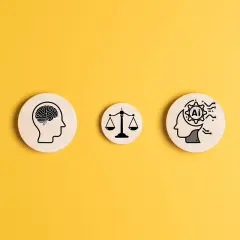 Trzy drewniane krążki na żółtym tle: ludzki mózg, waga sprawiedliwości i głowa z symbolem AI w środku, ilustrują, jak sztuczna inteligencja może rozwiązać dylematy etyczne.