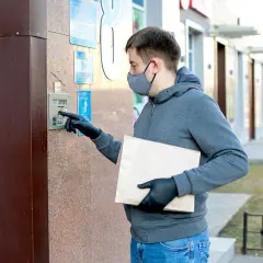 Mężczyzna w masce i rękawiczkach trzyma paczkę i wpisuje kod do domofonu, aby dostarczyć przesyłkę.