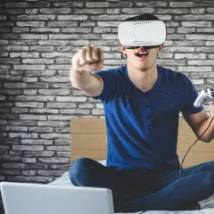 Chłopak w goglach VR z kontrolerem w ręku, siedzi na łóżku przed laptopem. Dowiedz się, jak podłączyć PS VR do PC i zanurz się w wirtualnym świecie!