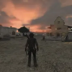 Postać w kapeluszu i z karabinem na plecach stoi na pustkowiu miasteczka o zachodzie słońca. Red Dead Redemption 2 PS Plus.