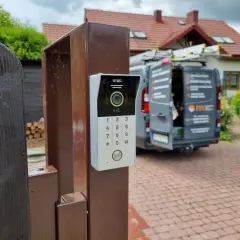 Nowy domofon na furtce. Zastanawiasz się, ile kosztuje montaż domofonu w domu jednorodzinnym? W tle widać dom i samochód firmy instalacyjnej.