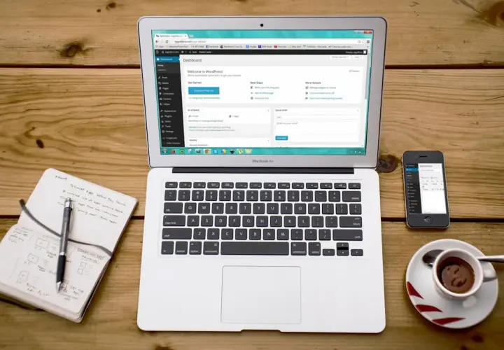 Laptop z panelem WordPress, notatnik z długopisem, telefon i filiżanka kawy. Idealne miejsce do nauki, jak zainstalować WordPress na komputerze.