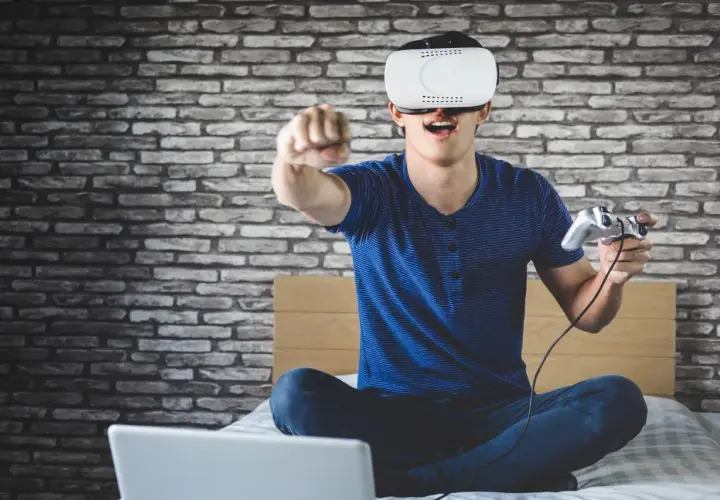 Chłopak w goglach VR z kontrolerem w ręku, siedzi na łóżku przed laptopem. Dowiedz się, jak podłączyć PS VR do PC i zanurz się w wirtualnym świecie!