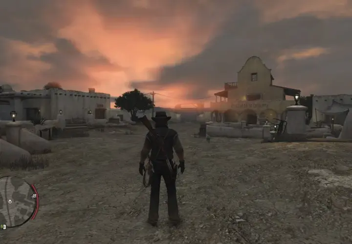 Postać w kapeluszu i z karabinem na plecach stoi na pustkowiu miasteczka o zachodzie słońca. Red Dead Redemption 2 PS Plus.