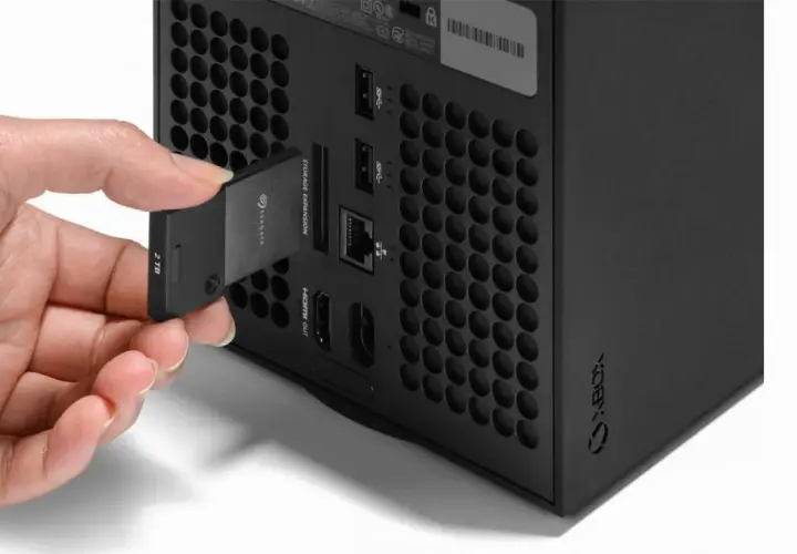 Dłoń wkłada kartę pamięci 2TB do dedykowanego portu rozszerzeń konsoli Xbox Series X, obok innych portów.