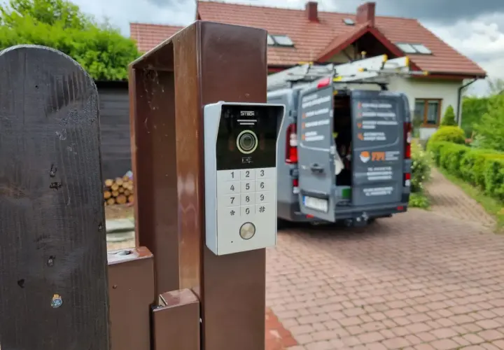 Nowy domofon na furtce. Zastanawiasz się, ile kosztuje montaż domofonu w domu jednorodzinnym? W tle widać dom i samochód firmy instalacyjnej.