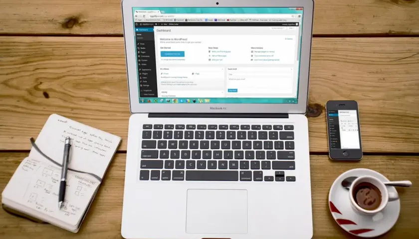 Laptop z panelem WordPress, notatnik z długopisem, telefon i filiżanka kawy. Idealne miejsce do nauki, jak zainstalować WordPress na komputerze.