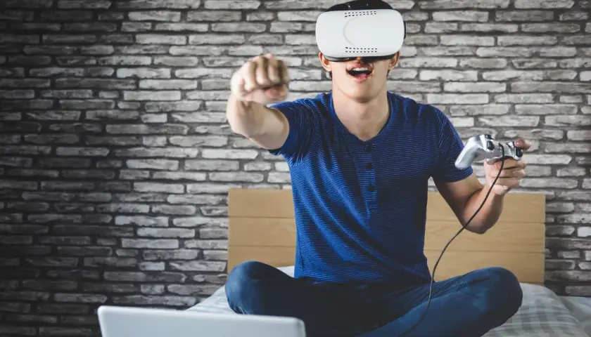 Chłopak w goglach VR z kontrolerem w ręku, siedzi na łóżku przed laptopem. Dowiedz się, jak podłączyć PS VR do PC i zanurz się w wirtualnym świecie!