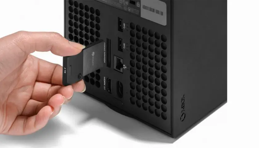 Dłoń wkłada kartę pamięci 2TB do dedykowanego portu rozszerzeń konsoli Xbox Series X, obok innych portów.