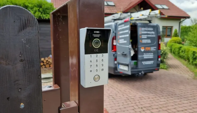 Nowy domofon na furtce. Zastanawiasz się, ile kosztuje montaż domofonu w domu jednorodzinnym? W tle widać dom i samochód firmy instalacyjnej.
