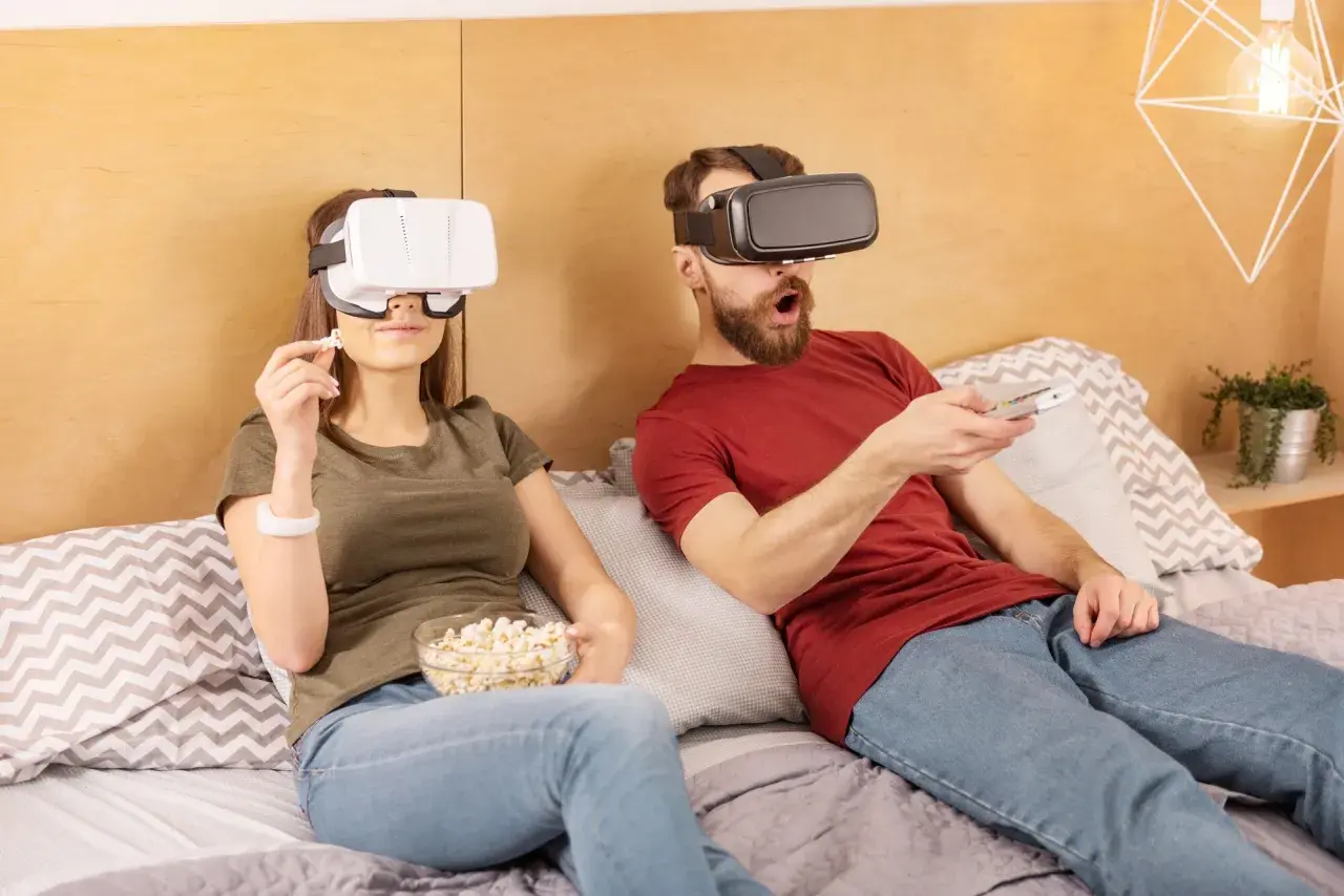 Para jak oglądać filmy VR na komputerze: para w goglach VR leży w łóżku, jedząc popcorn i trzymając pilota.