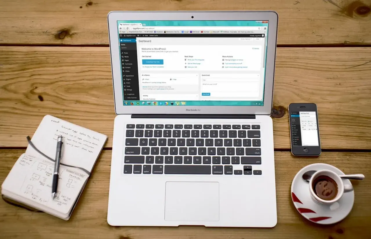 Laptop z panelem WordPress, notatnik z długopisem, telefon i filiżanka kawy. Idealne miejsce do nauki, jak zainstalować WordPress na komputerze.