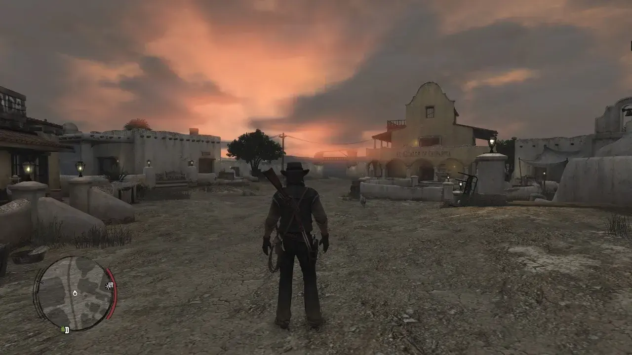 Postać w kapeluszu i z karabinem na plecach stoi na pustkowiu miasteczka o zachodzie słońca. Red Dead Redemption 2 PS Plus.