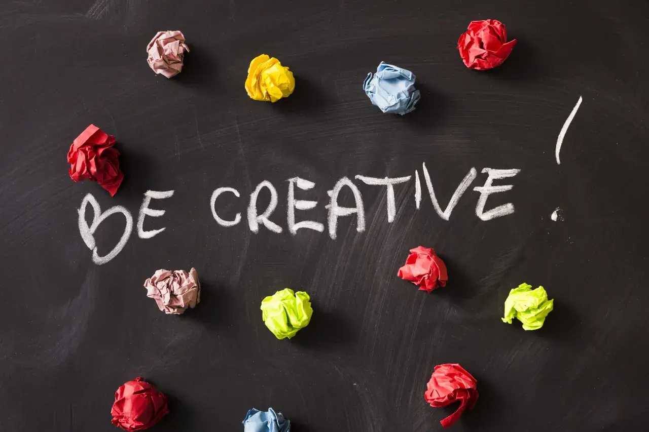 Kolorowe kulki papieru na czarnej tablicy, z napisem "BE CREATIVE!". To inspiracja, jak oszukać system binarny, myśląc nieszablonowo.