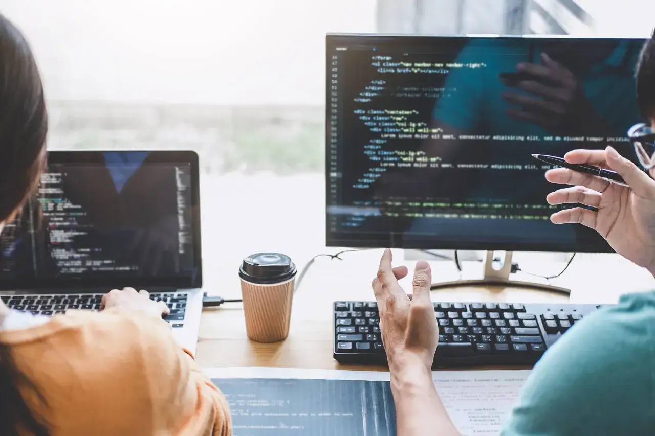 Dwóch programistów przy biurku, jeden pisze kod na laptopie, drugi wskazuje na ekranie monitora, omawiając jak pogrubić tekst w HTML.